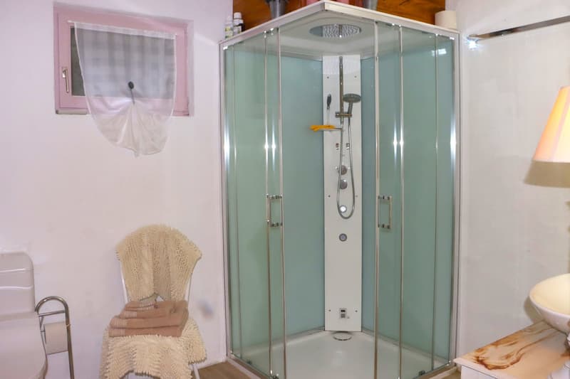 glass walk-in shower, white sink, towel on chair, towel hanging on rod, toilet, lamp