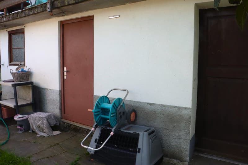 A house with a balcony above, white painted wall, brown door, garden hose reel, small table with a basket, doormat and a green hose.