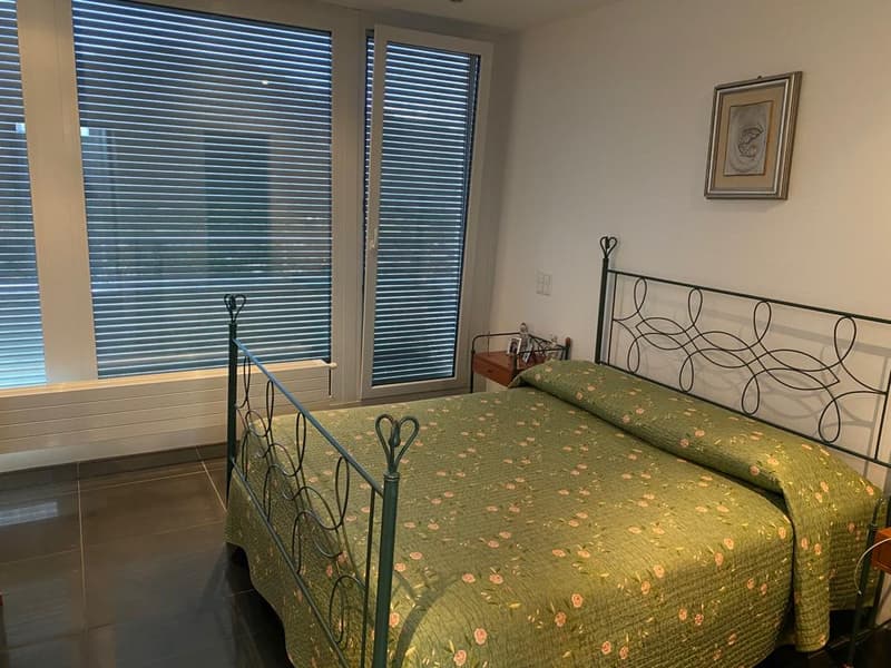 Single bed with green bedsheet, decorative pillows, nightstand with picture frame, white walls, white blinds
