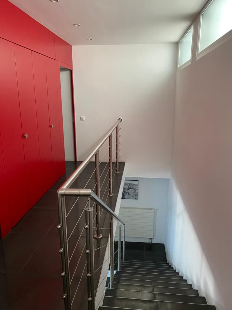 Modern staircase with metal handrail and tiled floor. Red cabinets on the left. Windows on the wall.