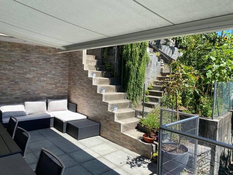 Furnished patio with a sofa, two stools, outdoor dining table, metal railing, outdoor stairs, planter with green plants, tiled wall, garden with various plants and sunflowers