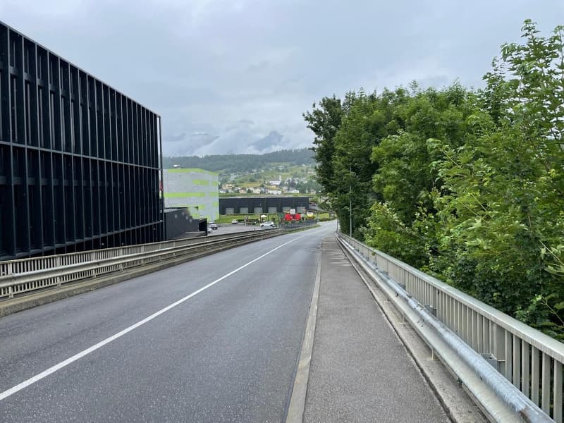 Empty road with steel guard rail, trees on one side, black structure on the other, cloudy sky, mountains in the background.