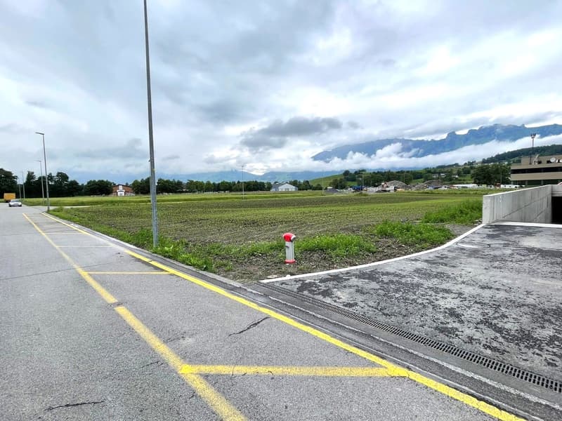 Exterior shot of a construction site, featuring an empty paved road, yellow markings, a pole with a fire extinguisher, a large grassy field, distant houses, and a building under construction, surrounded by cloudy sky and mountains.