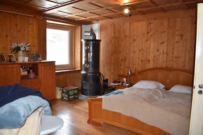 wooden walls, bed with two pillows, flower vase, lamp, wood-burning stove, cabinet with picture frames, desk with laptop