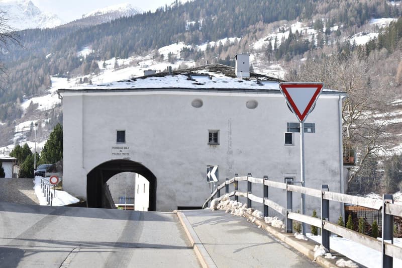 A white stone building, a tunnel entrance, snow-covered mountain, wooden fence, two traffic signs, bare trees