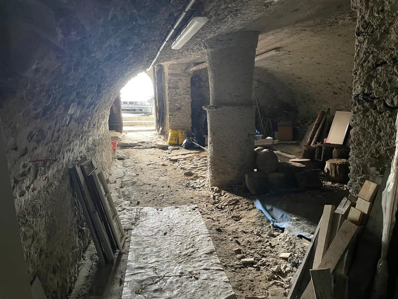 Archway tunnel with stone walls, beams, open area, light fixtures, doorway with car, yellow sacks, wooden pallets, debris, wooden posts