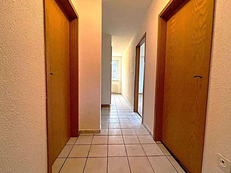 Empty hallway with tiled floor, wooden doors, and a window in the distance.