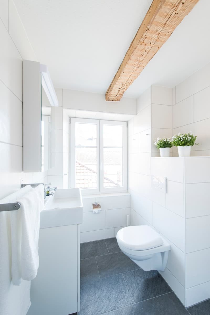 White walls, wooden ceiling beam, white vanity with sink, white toilet, gray tile floor, window