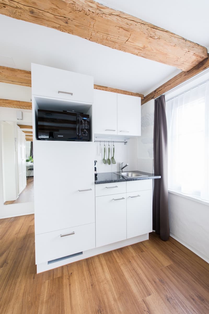 Compact kitchen with white cabinets, black microwave, wooden ceiling beams, and hardwood flooring.