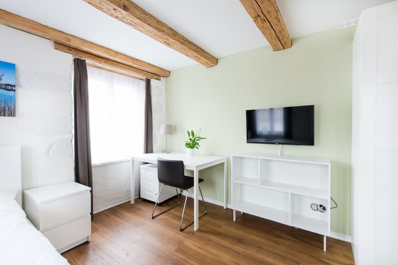 Spacious bedroom with wooden beams on the ceiling, large window, white walls, wooden floor, desk with chair, TV stand, and a potted plant