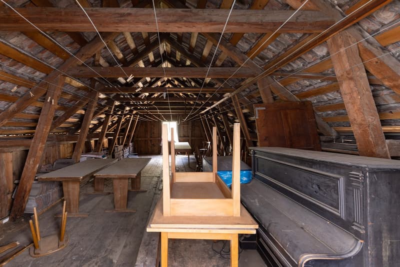 The image shows an old, wooden attic space with a high, slanted ceiling and exposed wooden beams. The attic appears to be used for storage, with various items and furniture visible, including wooden tables and chairs, and a large wooden chest or cabinet.