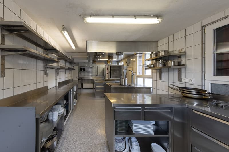 Large commercial kitchen with stainless steel appliances, shelves, and countertops. The kitchen appears to be well-equipped with various cooking equipment and storage for dishes and utensils.