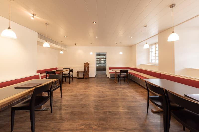 Large dining room with wooden floors, red upholstered benches along the walls, wooden tables and chairs, pendant lighting fixtures, and a window at the back of the room.