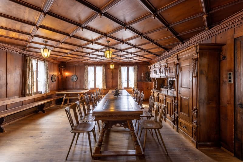 Large wooden dining room with a high, ornate wooden ceiling, wooden walls, and a long wooden dining table with chairs. The room features a fireplace and decorative wall sconces providing warm lighting.