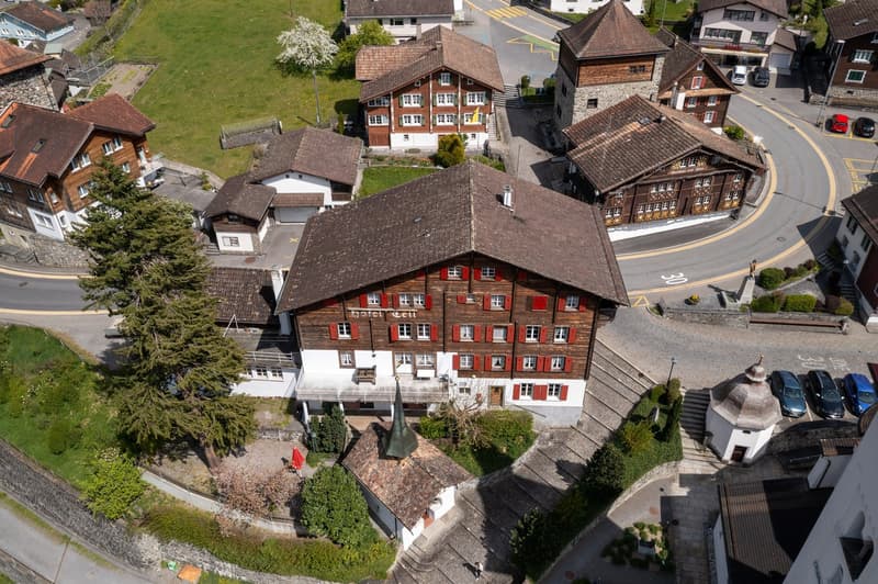 A traditional Swiss-style hotel building with a red-tiled roof, surrounded by other residential and commercial buildings in a small town. The hotel has a courtyard and garden area, and there are parking spaces visible in front of the building.