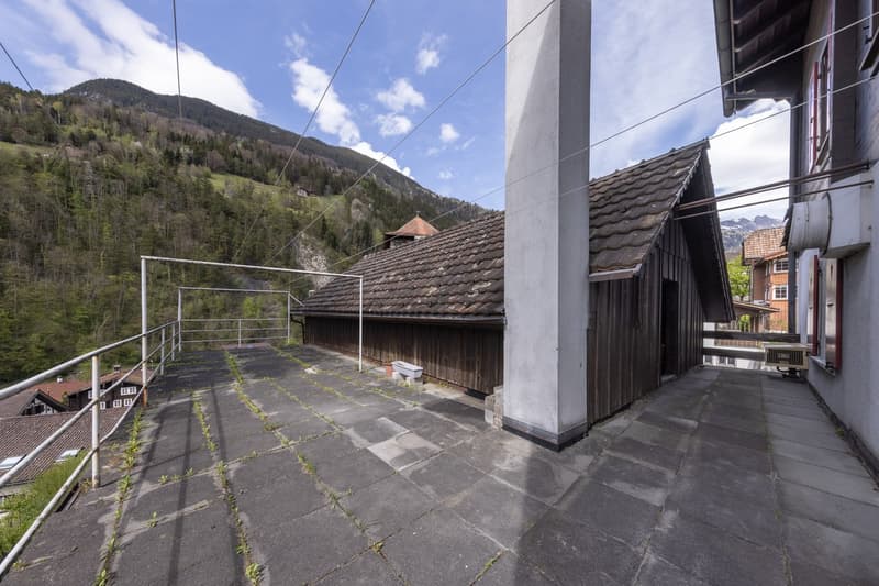 The image shows the exterior of a building with a balcony or terrace. The building has a slate roof and wooden siding. There is a concrete walkway leading to the building, and the surrounding landscape includes mountains and forested hills.