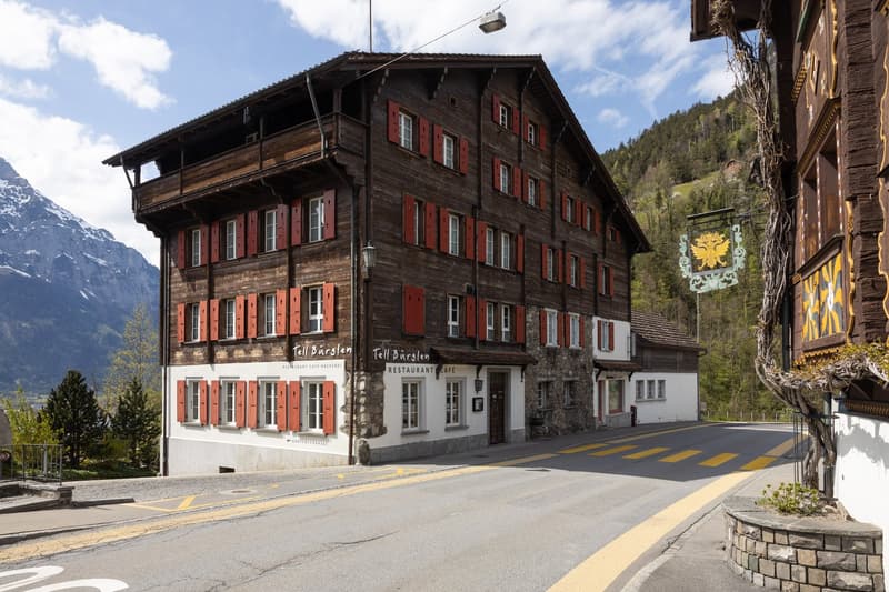 A traditional Swiss chalet-style building with wooden exterior and red shutters. The building has a restaurant and cafe on the ground floor, and appears to be located in a mountainous, scenic area with a road passing in front of it.
