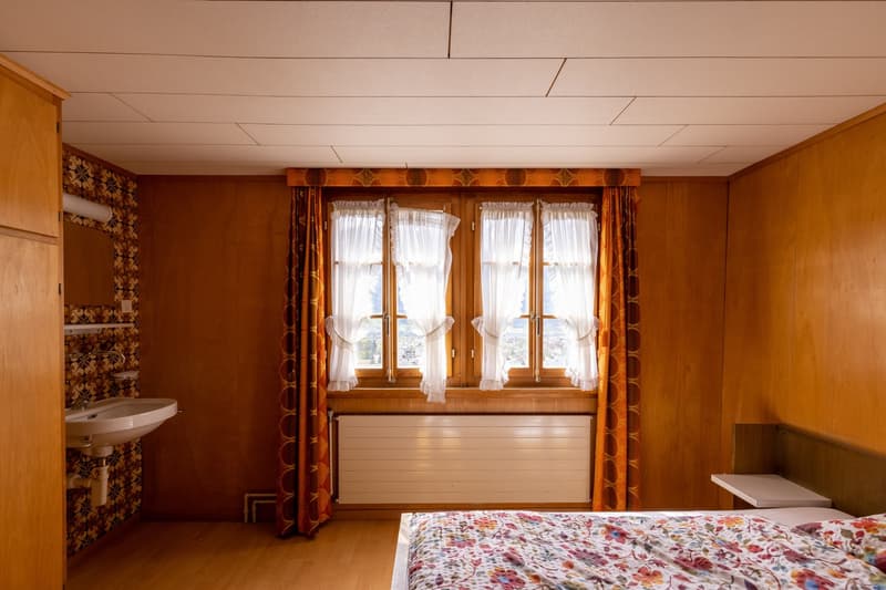 Cozy bedroom with wood-paneled walls, large windows, floral curtains, and a floral bedspread. There appears to be a balcony or terrace visible through the windows.
