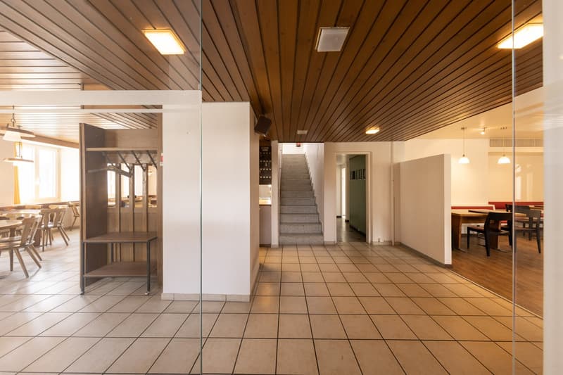 The image shows the interior of a building with a wooden vaulted ceiling, tile flooring, and a glass partition wall. There is a staircase leading to an upper level, and the space appears to be divided into different areas, including a dining area with tab