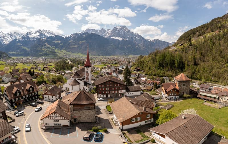 Picturesque alpine town with traditional Swiss-style buildings, a church steeple, and surrounded by snow-capped mountains and lush green hills. The town appears to have a mix of residential and commercial buildings, with cars parked on the streets.