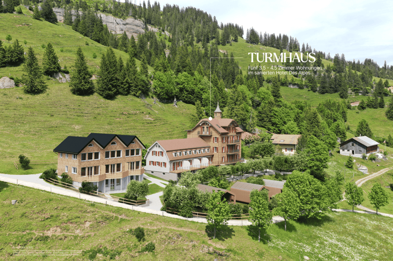 A multi-story hotel building with a traditional alpine architecture, surrounded by lush green hills and forests. The hotel appears to have balconies and terraces, offering scenic views of the natural landscape.