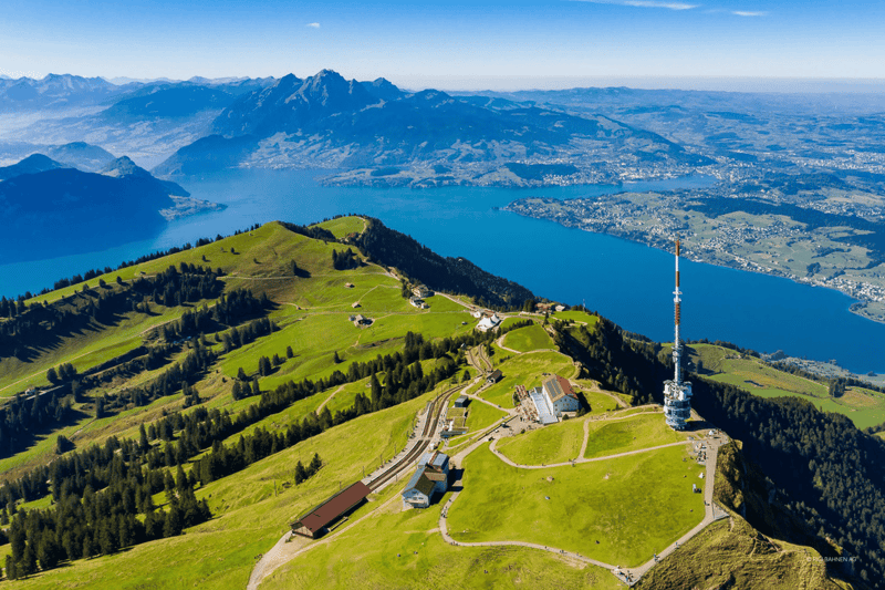 Aerial view of a mountainous landscape with a large lake, green hills and valleys, and a tall communication tower on the top of a hill. The scene offers a panoramic view of the surrounding mountains and towns.