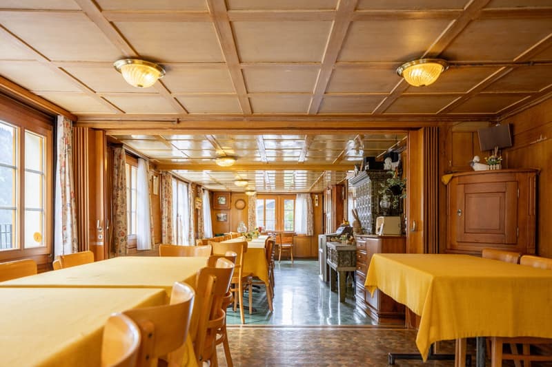 Spacious dining room with wooden paneled ceiling, large windows, wooden furniture including tables and chairs, and a fireplace visible in the background.