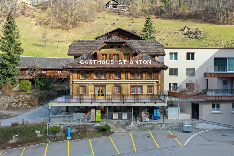 A traditional Swiss-style wooden building with a large sign that says 'Gasthaus St. Anton'. The building has a sloped roof and colorful window frames. There is a parking area in front of the restaurant.