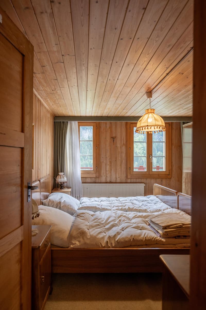Cozy wooden bedroom with a large bed, wooden walls and ceiling, and a decorative lamp fixture. The room has a warm, rustic feel with a fireplace visible.