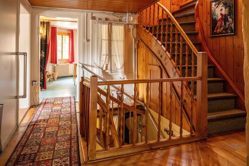 Rustic wooden staircase with ornate railing, hardwood floors, patterned area rug, curtained windows, and a framed artwork on the wall