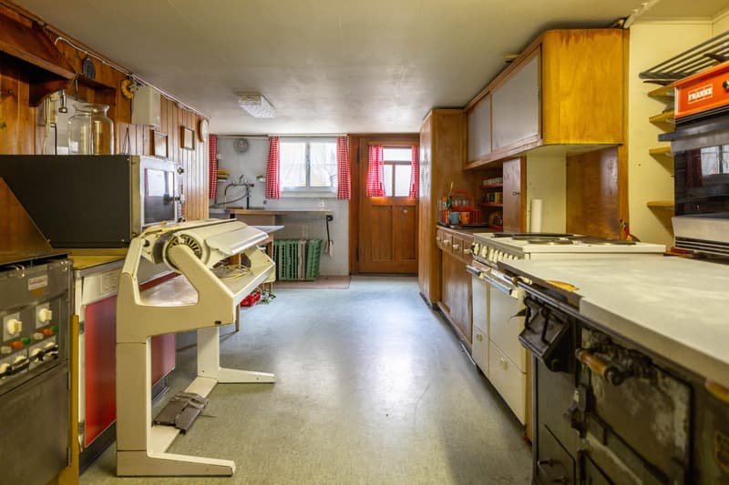 Vintage kitchen with wooden cabinets, old appliances, and a washing machine. The room has a cluttered and lived-in feel with various items and tools scattered around.