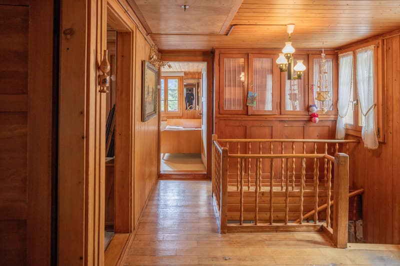Cozy wooden interior with a hallway, wooden floors, paneled walls, and a wooden staircase. The room features a fireplace and decorative elements like curtains and wall art. The overall atmosphere is warm and rustic.