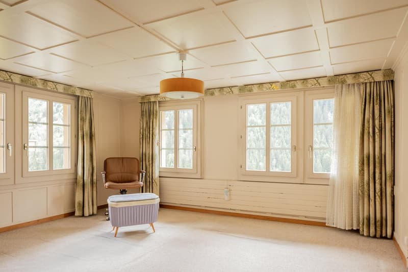 Spacious living room with large windows, wooden paneled walls, beige curtains, a brown leather armchair, and a storage ottoman.
