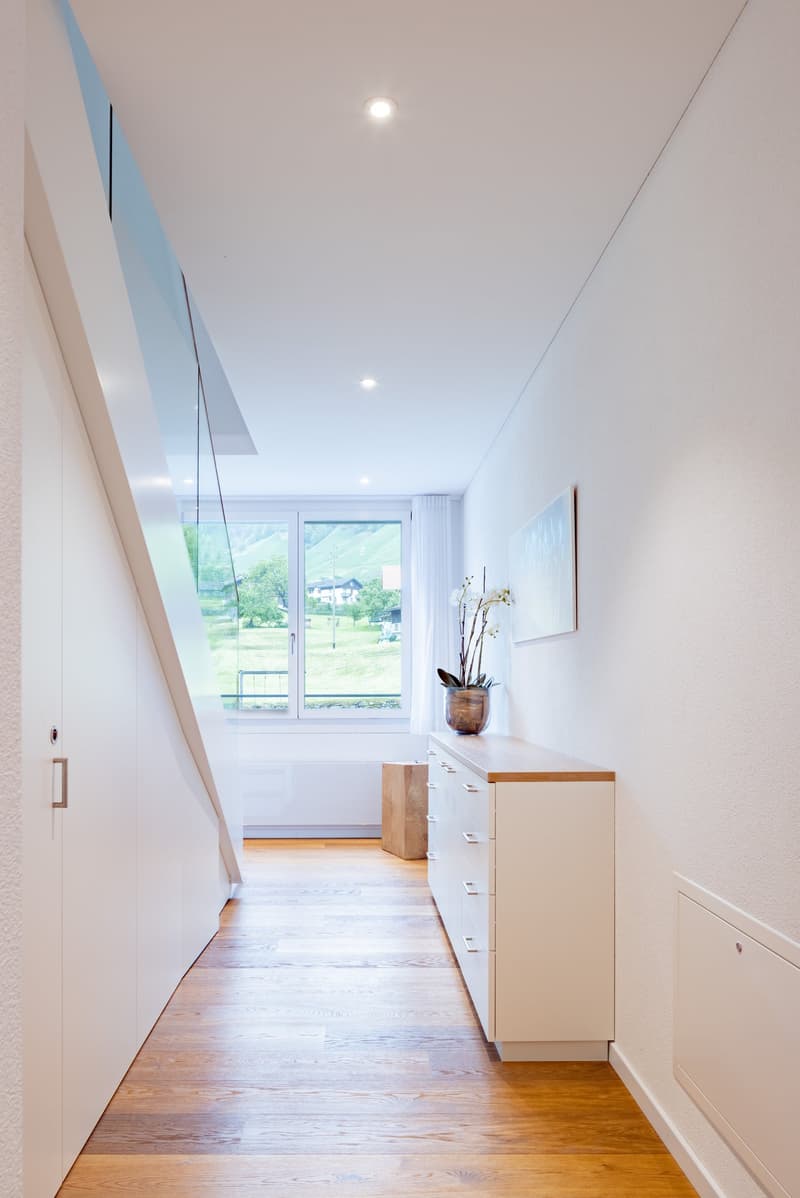 Bright and spacious hallway with large windows overlooking a scenic outdoor view. The hallway features hardwood floors, white walls, and built-in cabinets and drawers. A decorative vase with flowers is placed on a wooden shelf, adding a touch of nature to