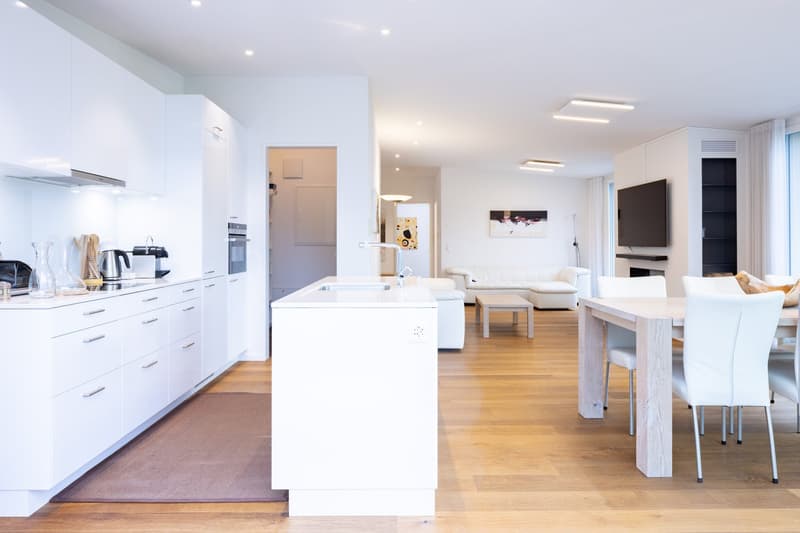 The image shows an open concept living space with a kitchen, dining area, and living room. The kitchen features white cabinets and countertops, with a dishwasher visible. The dining area has a wooden table and chairs, while the living room has a white sof
