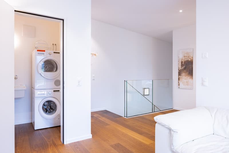 The image shows a room with a stacked washer and dryer unit, hardwood floors, and a glass railing leading to an upper level. The room appears to be a laundry or utility room with the washing machine and dryer visible.