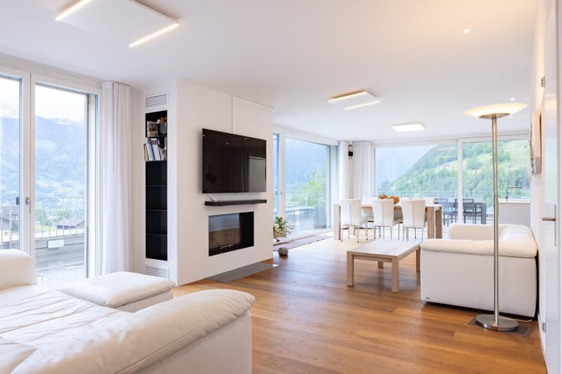 Spacious open-plan living room with large windows offering a scenic mountain view, hardwood flooring, white walls, modern furniture including a sofa, coffee table, and dining table with chairs, a wall-mounted TV with a fireplace below, and recessed lighti