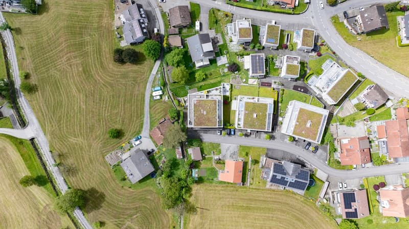 This image shows an aerial view of a residential area with a mix of houses, apartments, and other buildings. The area has a combination of green spaces, such as lawns and gardens, as well as paved areas for parking. Some of the buildings appear to have so