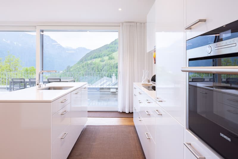 Spacious modern kitchen with large windows overlooking a scenic mountain landscape, white cabinets and countertops, stainless steel appliances including an oven, and a balcony or terrace area