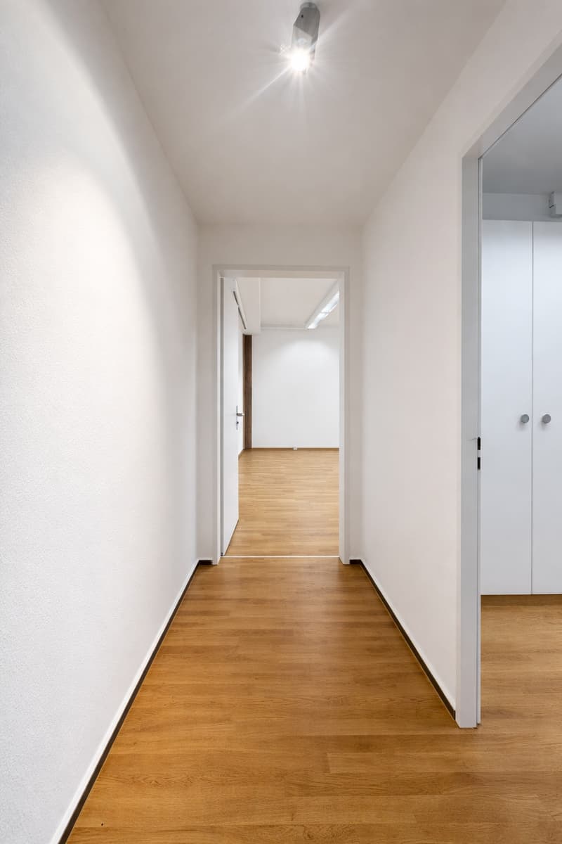 Empty hallway with wooden floor, white walls, and ceiling lights