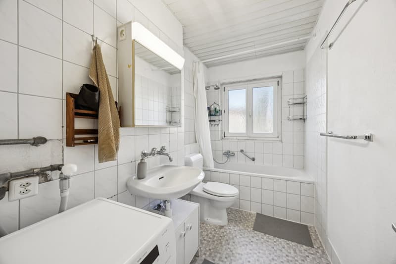 White tiled walls, white toilet, white bathtub, white sink, mirror, towel rack, power outlet, washing machine