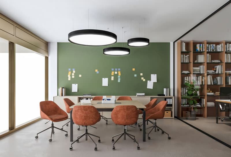 modern office room with round table, orange swivel chairs, green wall with sticky notes, bookshelves, and ceiling lights
