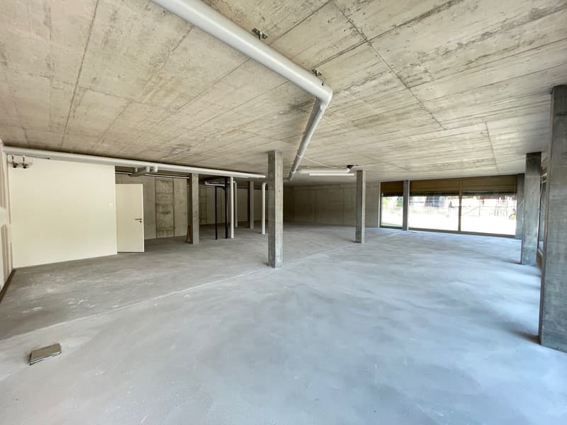 Empty room with concrete floor, white walls, large windows, and exposed concrete ceiling with pipes
