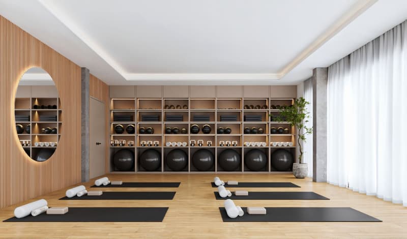 Yoga studio with wooden floor, white walls, shelves with exercise equipment, yoga mats, and a plant