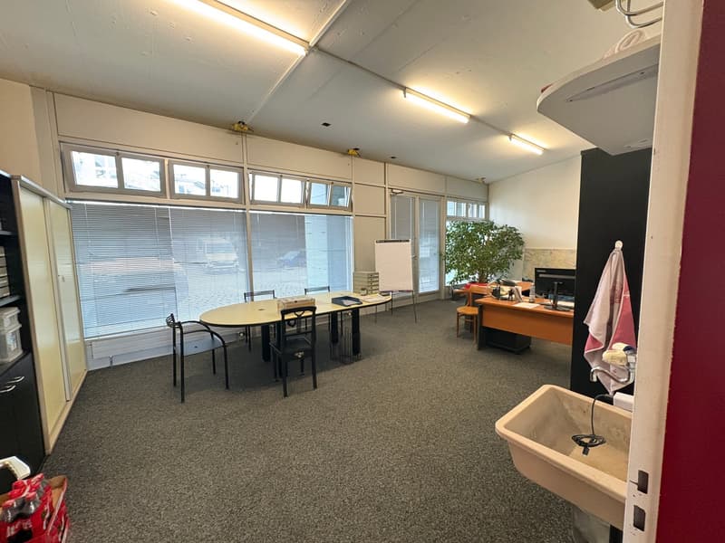 A large rectangular office space with white walls and gray floor carpet, with office chairs and tables, an oak desk, and a potted plant near the window, as well as fluorescent lights on the ceiling.