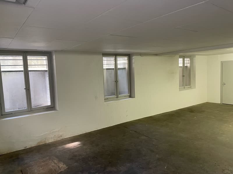 The image shows an empty room with white walls, a concrete floor, and several windows. The room appears to be in a state of renovation or preparation for a new use.