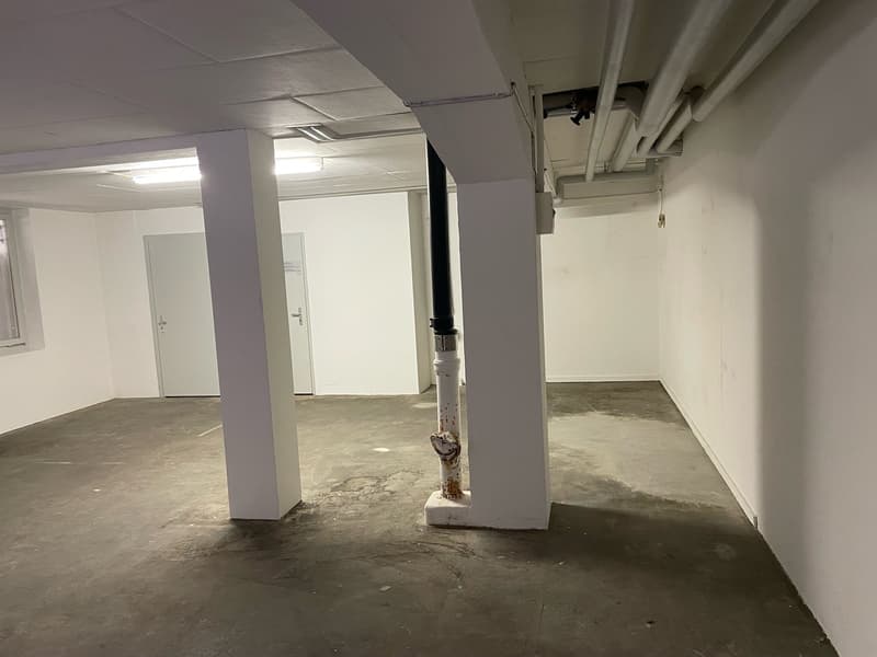 The image shows an empty office space with white walls, a concrete floor, and exposed ceiling pipes. The space appears to be in the process of renovation or construction, with a few doors visible in the background. The overall impression is of a bare, unf