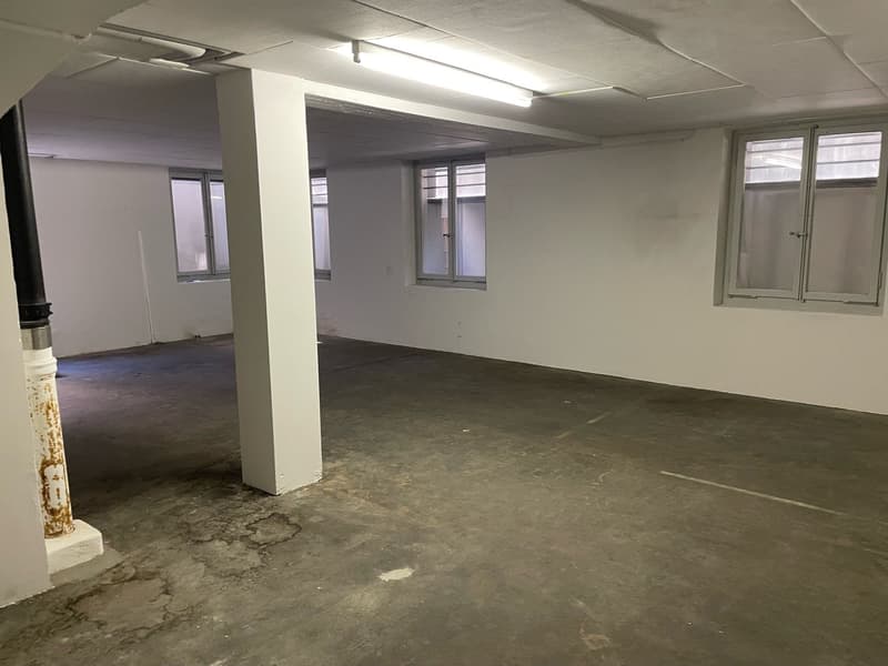 The image shows a large, open office space with concrete flooring, white walls, and overhead lighting. The space appears to be unfinished or in need of renovation, with some visible damage or wear on the floor. There are windows along one wall, providing