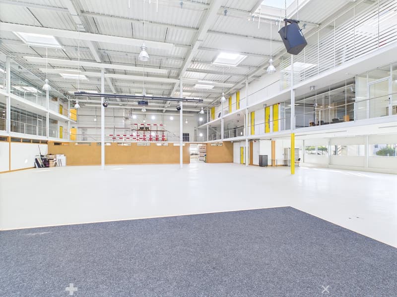 Large open space with high ceilings, concrete floor, and glass walls. The space appears to be a warehouse or industrial facility with ample lighting and storage areas.