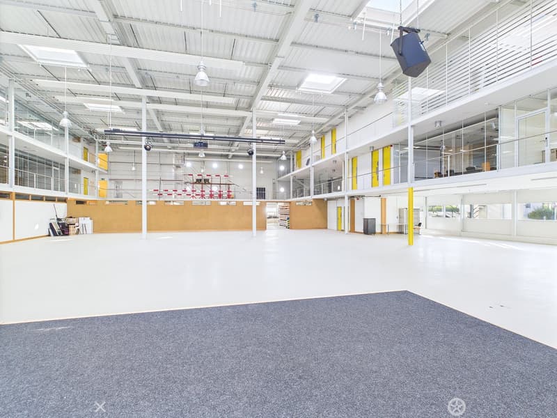 Large open space with high ceilings, concrete floor, and glass walls. The space appears to be a warehouse or industrial facility with ample lighting and storage areas.